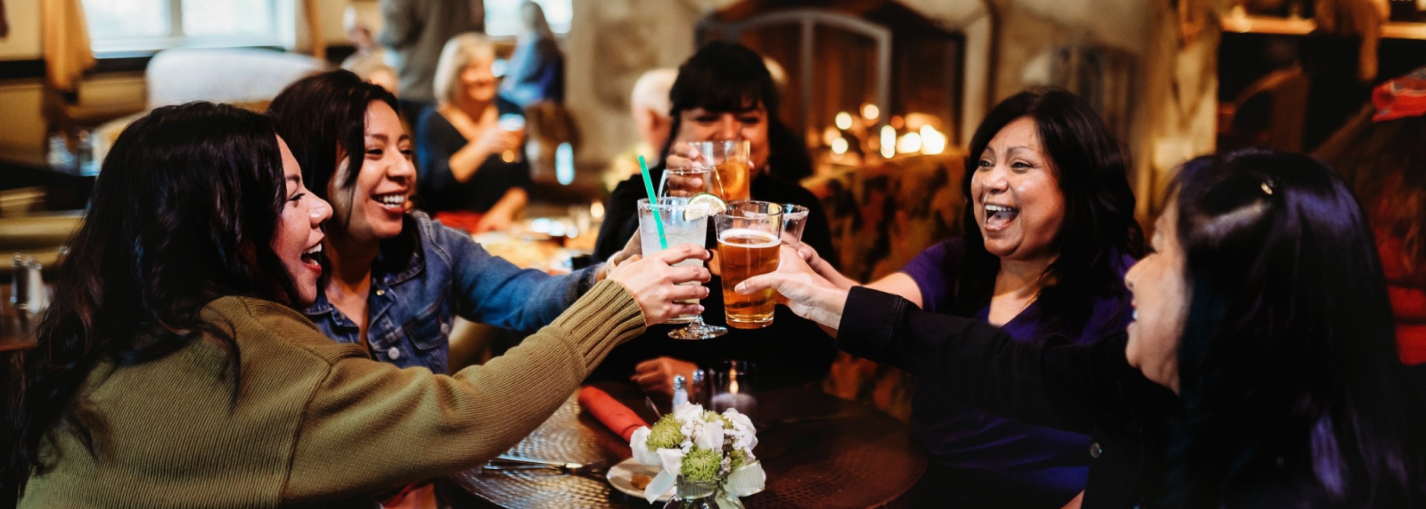 Group of women smiling, clinking drinks in front of fireplace at Fireside Lounge at the Oregon Garden Resort